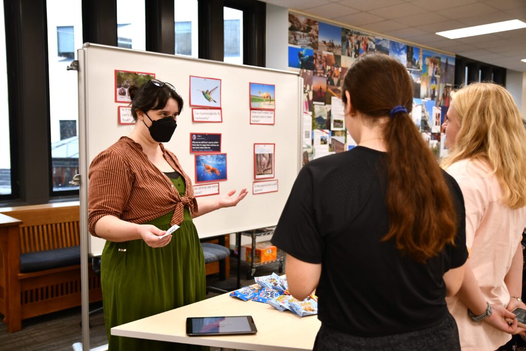 Photo shows a librarian talking to students at a table in the library with pictures for evaluation posted behind the librarian.